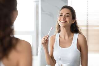 young female cleaning and brushing teeth with toothbrush standing near mirror